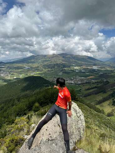 Ein Mann in Sportkleidung sitzt auf einem großen Felsen auf einem Hügel und zeigt auf ein malerisches Tal mit grünen Wiesen, Wäldern und Bergen unter dramatischen Wolken.
