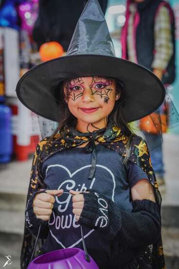 Ein junges Mädchen, das sich zu Halloween als Hexe verkleidet hat: Sie trägt einen schwarzen Hut und einen Umhang, hat Spinnennetz-Gesichtsbemalung, hält einen lila Kürbiskübel in der Hand und lächelt selbstbewusst.