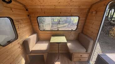 Cette photo montre l'intérieur chaleureux d'un camping-car en bois, avec deux banquettes rembourrées, une petite table rectangulaire entre les deux et de grandes fenêtres laissant entrer beaucoup de lumière naturelle.