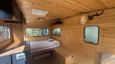 Cette image montre l'intérieur chaleureux d'un camping-car, avec des boiseries, un lit, un bureau, des fenêtres, une étagère en hauteur, des bois de cerf et un chapeau accroché au mur.