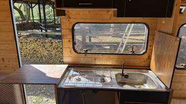Cette photo montre une cuisine de camping-car compacte équipée d'une cuisinière à deux feux, d'un petit évier, d'un plan de travail en bois, d'armoires suspendues et d'une grande fenêtre donnant sur un tas de bois de chauffage.