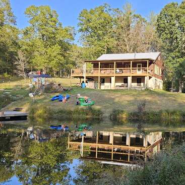 A rustic lakeside cabin sits on a grassy hill, surrounded by trees. Colorful kayaks and toys lay outside, reflecting beautifully in the calm water below.