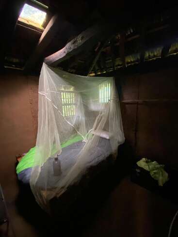 A small, rustic room with a bed covered by a mosquito net, dim natural light entering from a window and skylight, creating a cozy atmosphere.