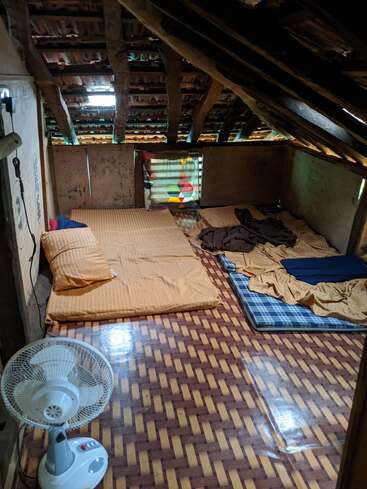 This image shows a rustic attic room with a wooden roof, patterned floor, simple bedding on the floor, a colorful curtain, and a standing electric fan.