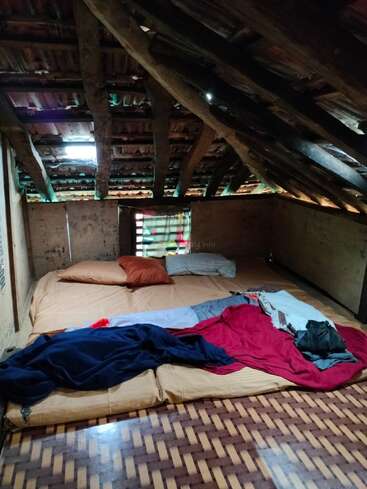 This cozy attic room features a slanted wooden roof, two pillows, a mattress with blankets, dim light filtering through gaps, and rustic, earthy walls.