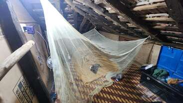 A rustic attic room with a slanted wooden roof, features a bed covered by a mosquito net. There are bags, a blue chest, and exposed beams.