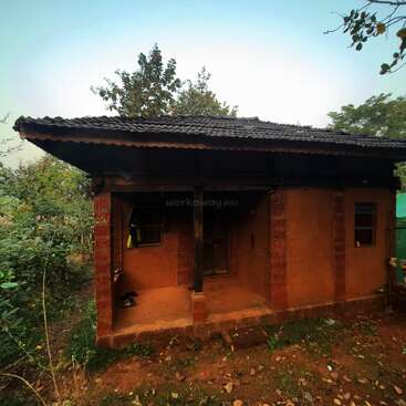 This image shows a small, rustic house with mud walls and a tiled roof, surrounded by greenery and trees, exuding a peaceful, rural atmosphere.