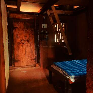 This rustic room features earthy walls, a wooden door, a blue checkered mattress on a wooden bed, a loft with wooden stairs, and minimal natural lighting.