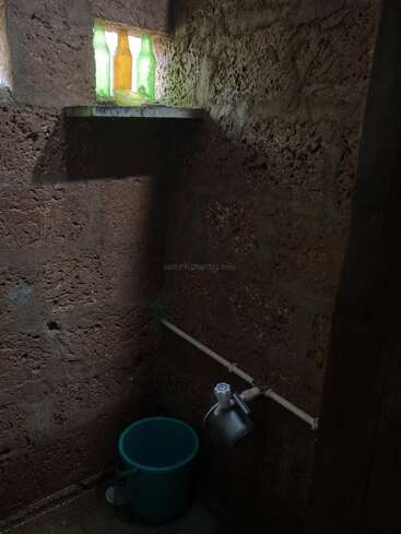 A rustic bathroom with rough textured walls, a green bucket, a metal mug, a tap, and a shelf with three colored bottles lit by sunlight.