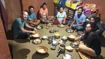 A group of eight people sit on the floor in a cozy room, sharing a traditional Indian meal served in metal plates and bowls, smiling together warmly.