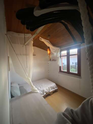 A small, cozy bedroom with wooden ceiling, simple white bedding, pallet bed, large window, soft natural light, and decorative hanging white fabric creating a peaceful atmosphere.