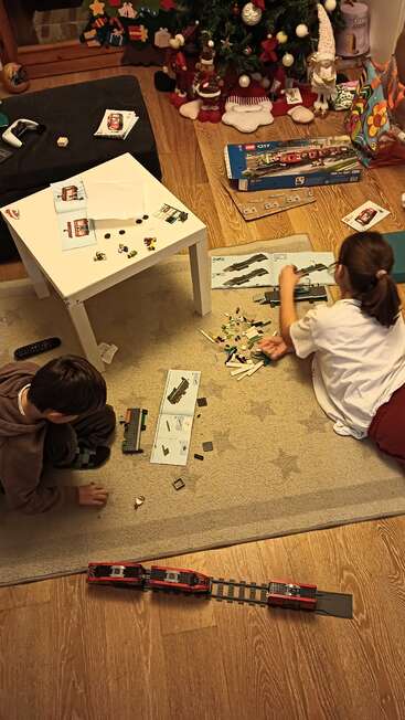 Zwei Kinder bauen auf einem Teppich eine LEGO-Eisenbahn zusammen. Weihnachtsdeko und ein Weihnachtsbaum stehen in der Nähe und sorgen für eine fröhliche und verspielte Atmosphäre im Haus.