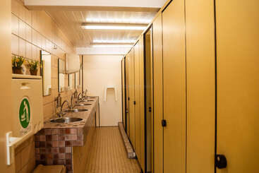 Esta imagen muestra un baño público limpio con puertas amarillas en los cubículos, varios lavabos con espejos, paredes alicatadas en color beige, un cambiador para bebés e iluminación fluorescente.