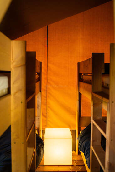 Esta imagen muestra una acogedora habitación con literas de madera a ambos lados, una pared revestida de tela naranja y una lámpara luminosa con forma de cubo que proporciona una iluminación cálida y ambiental en el centro.