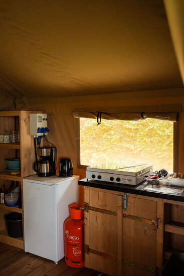 Una acogedora cocina de campamento con una nevera pequeña, una cafetera, un hervidor, una cocina de gas, estantes de madera, vajilla, utensilios y una bombona de gas naranja debajo de una ventana.