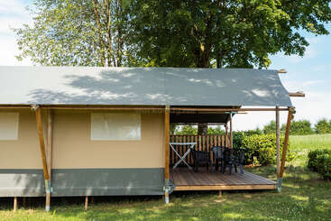 En esta imagen se ve una moderna tienda de glamping situada en una zona verde y cubierta de césped, con una terraza de madera cubierta, sillas de plástico, una mesa y árboles altos que proporcionan sombra.