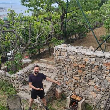 The image depicts a man standing in a garden, gesturing with his hands, surrounded by a stone wall, trees, and a table with food and drink items.
