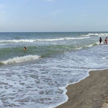 Das Bild zeigt eine ruhige Strandszene mit sanften Wellen, die an das Ufer plätschern, und drei Personen, die an einem sonnigen Tag am Wasser entlang spazieren.