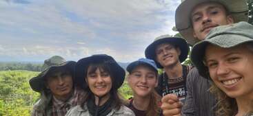 Sechs Menschen mit Hüten posieren für ein fröhliches Selfie im Freien, mit üppiger grüner Vegetation und einem wolkenverhangenen Himmel im Hintergrund, der vermuten lässt, dass sie auf einem Abenteuer sind.