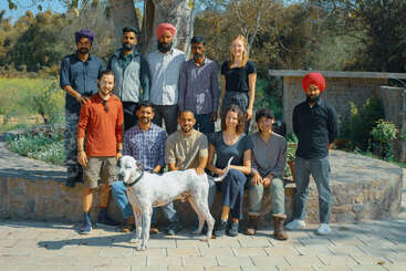 A diverse group of eleven people and a white dog pose outdoors on a sunny day. Some are sitting, some standing, with trees and greenery behind them.