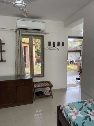 A neat, airy room with a window and door facing outside, air conditioner, wooden cabinet, shoe rack, bed with floral sheets, and wall hooks.