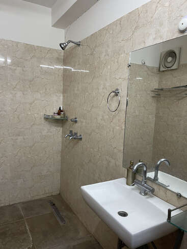 This is a modern bathroom with beige marble tiles, a wall-mounted shower, a large mirror, a white sink, a towel ring, and minimal toiletries.