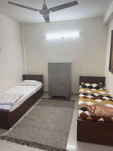 This image shows a simple, clean room with two single beds, a metal wardrobe, a ceiling fan, a rug, white walls, and bright lighting.