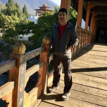 Un hombre de pie sobre un puente de madera, con las manos en los bolsillos, con árboles, montañas y un edificio tradicional butanés de fondo bajo la luz del sol.