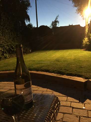 The image depicts a serene backyard scene at dusk, featuring a bottle of wine and a glass on a table, with a lush green lawn and trees in the background.