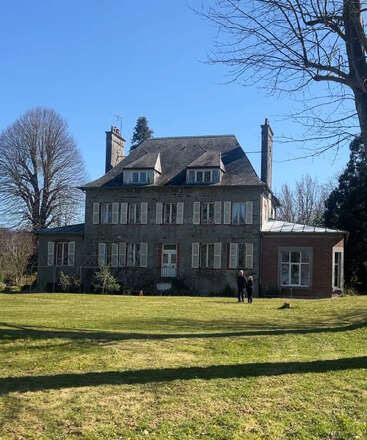 A large, old brick house with many windows sits in a spacious yard. Two people stand out front under a clear blue sky, surrounded by trees.