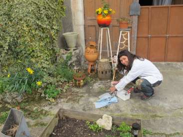 Das Bild zeigt eine Frau, die einen hölzernen Pflanzkasten in einem Garten bemalt, umgeben von verschiedenen Pflanzen und Dekorationsgegenständen.