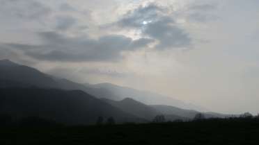 Das Bild zeigt eine Bergkette mit einem wolkenverhangenen Himmel und einer Sonne, die durch die Wolken blickt und einen dunstigen Schein über die Szene wirft.