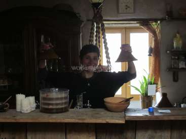 Das Bild zeigt einen Mann, der hinter einem hölzernen Tresen steht, einen Trichter in der Hand hält und einen großen Behälter mit Flüssigkeit sowie verschiedene Gegenstände auf dem Tresen stehen hat.