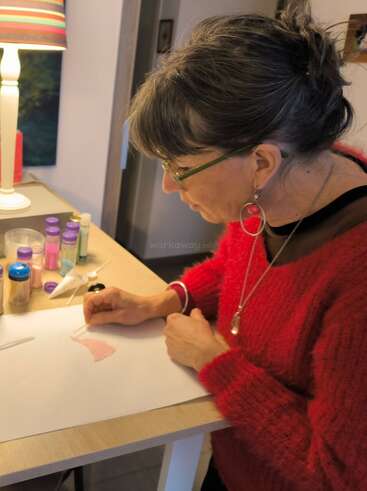 Une femme portant des lunettes et un pull-over rouge décore une feuille de papier à un bureau, entourée de bouteilles colorées, sous une lampe rayée en intérieur.