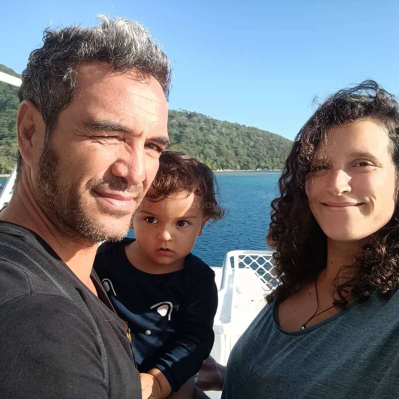 The image shows a family of three posing for a selfie on a boat, with a serene body of water and a lush green hillside in the background.