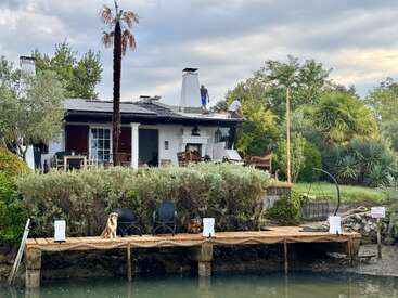 A white house with a porch sits by the water. Two people work on the roof. Dogs relax on the wooden dock amid lush greenery.