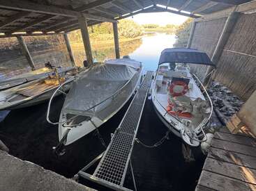 Under a wooden shelter, three boats are docked on calm water. One boat is covered, another has a canopy, surrounded by ropes, nets, and life preservers.