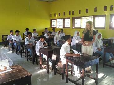 Une salle de classe aux murs jaunes, remplie d'élèves en uniforme blanc. Un enseignant se tient près du bureau d'un élève, interagissant avec lui, tandis que d'autres semblent attentifs et engagés.