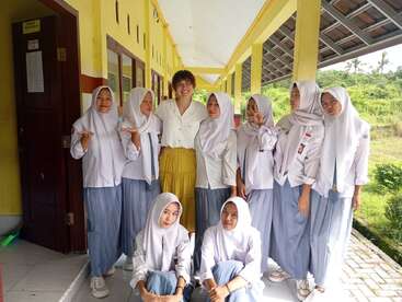 Un groupe de neuf filles portant des uniformes scolaires et des hijabs se tient aux côtés d'une femme souriante vêtue d'un pantalon jaune, posant joyeusement ensemble dans un couloir d'école.