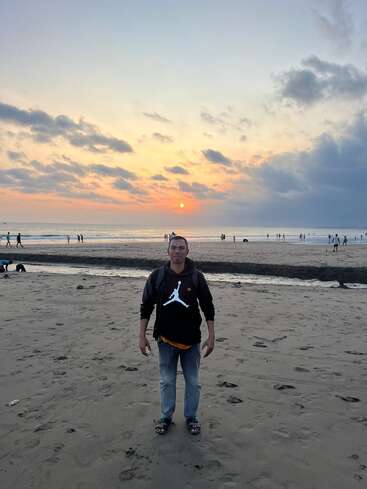Un homme se tient sur une plage de sable au coucher du soleil, en tenue décontractée. Le ciel est nuageux, le soleil se couche et les gens profitent de la sérénité du rivage.