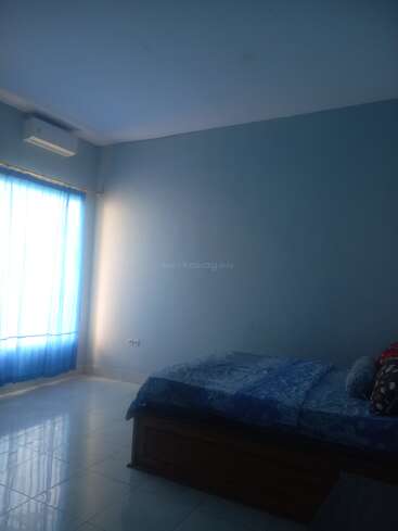 Cette image montre une chambre à coucher simple avec des murs bleus, un lit bien fait, des rideaux filtrant la lumière du soleil, un sol carrelé et un climatiseur au-dessus de la fenêtre. Décoration minimale.