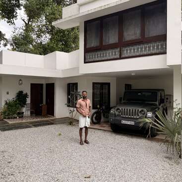 A man stands in front of a modern white house with large windows. A jeep and motorcycle are parked in the driveway, surrounded by plants and greenery.