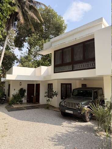 Una moderna casa blanca de dos plantas con grandes ventanales, rodeada de árboles y vegetación. Un Jeep está aparcado en el camino de entrada sobre grava, bajo un cielo soleado.