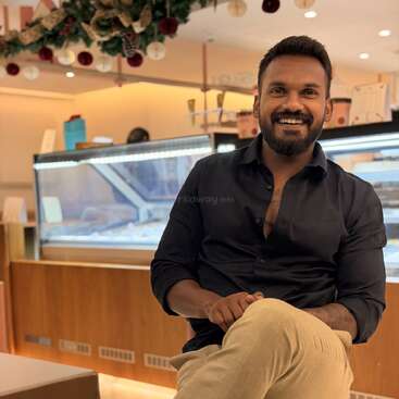 Ein lächelnder Mann sitzt gemütlich in einem gemütlich beleuchteten Café und trägt ein dunkles Hemd und eine helle Hose. Hinter ihm sieht man festliche Deko und Vitrinen.