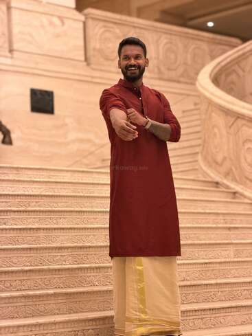 A smiling man in a maroon kurta and cream dhoti stands on ornate marble stairs, rolling up his sleeves, posing confidently in a grand, elegant setting.