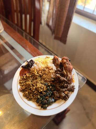 A white plate is filled with rice, leafy greens, lentils, chicken drumsticks, beef curry, and sauces, placed on a glass dining table near a window.