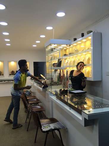 L'image représente un homme achetant des bijoux à une femme au comptoir d'un magasin, avec une vitrine de bijoux derrière eux et une rangée de chaises le long du mur.