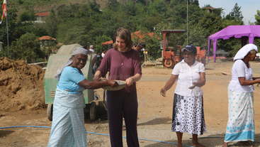 L'image montre un groupe de personnes réunies autour d'une table, avec quelques individus debout à l'arrière-plan. La scène semble être un rassemblement ou un événement communautaire, peut-être pour une occasion sociale ou culturelle.