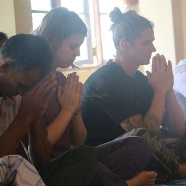 L'image représente trois personnes, les mains jointes devant elles, assises sur le sol, dans une pièce avec des fenêtres et un mur blanc.