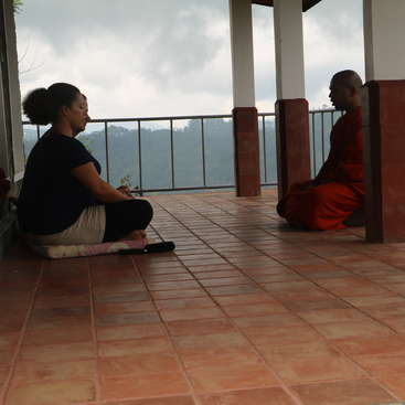 L'image représente deux personnes assises sur un sol carrelé, l'une face à l'autre, avec une balustrade et des montagnes visibles à l'arrière-plan, dans un ciel couvert.
